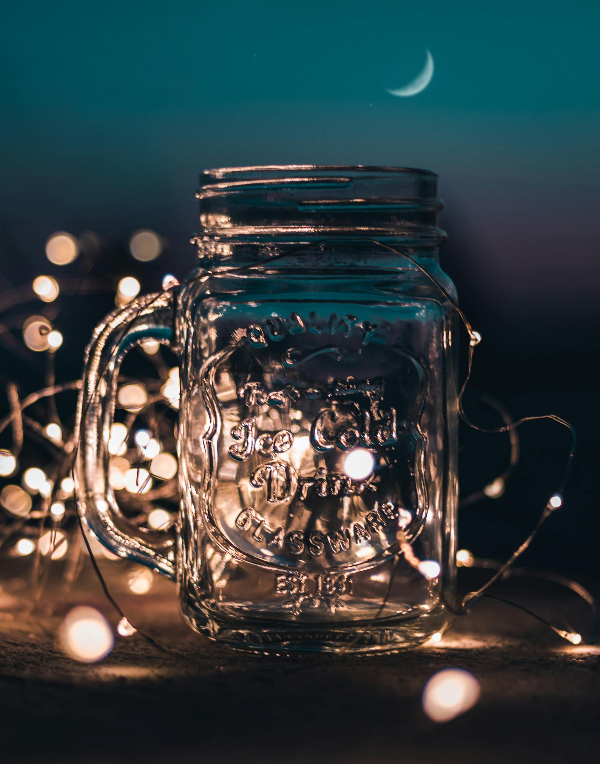 A clear mason jar illuminated by fairy lights set against a nighttime sky with a crescent moon.