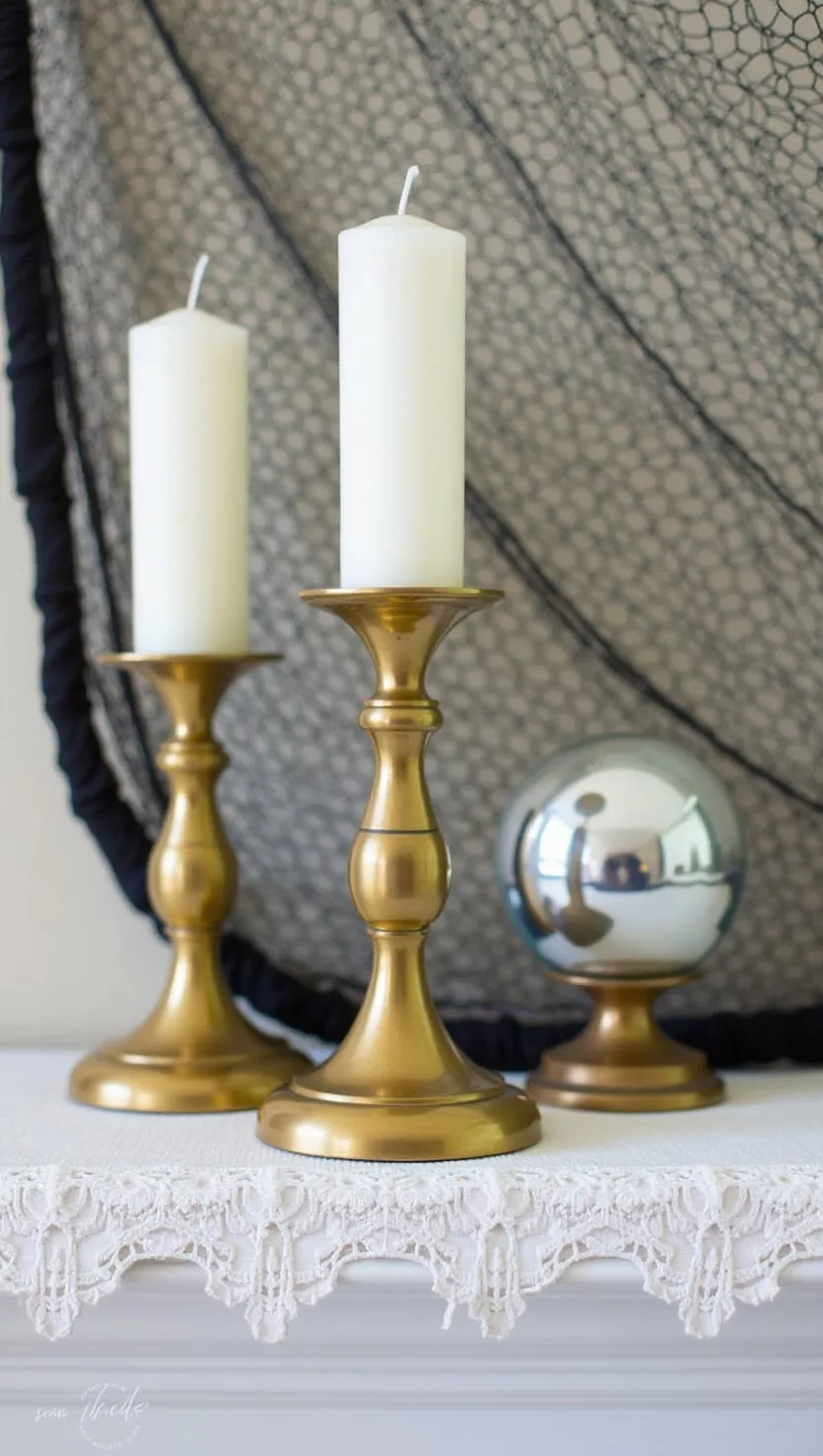 A photo of two brass candlesticks with white candles beside a mercury glass gazing ball on a lace-edged mantle, with a black fishing net draping behind.